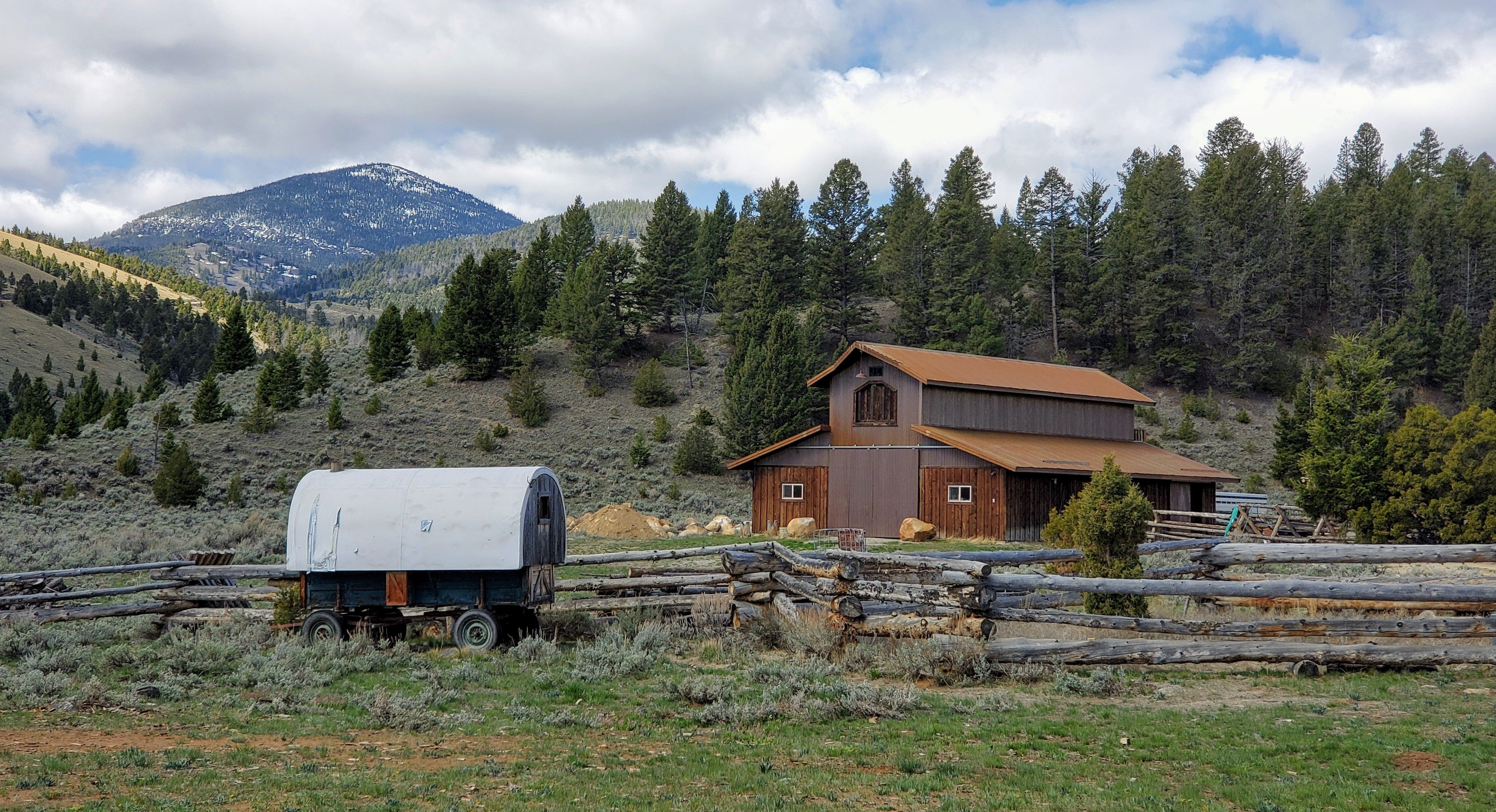 A photo of an old covered wagon next to a brown barn in a mountain valley