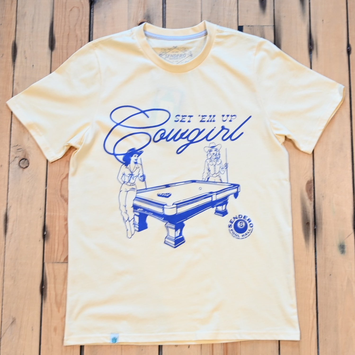 Unisex cream tee by Sendero with graphic of two cowgirls playing pool and “Set ’Em Up Cowgirl” text