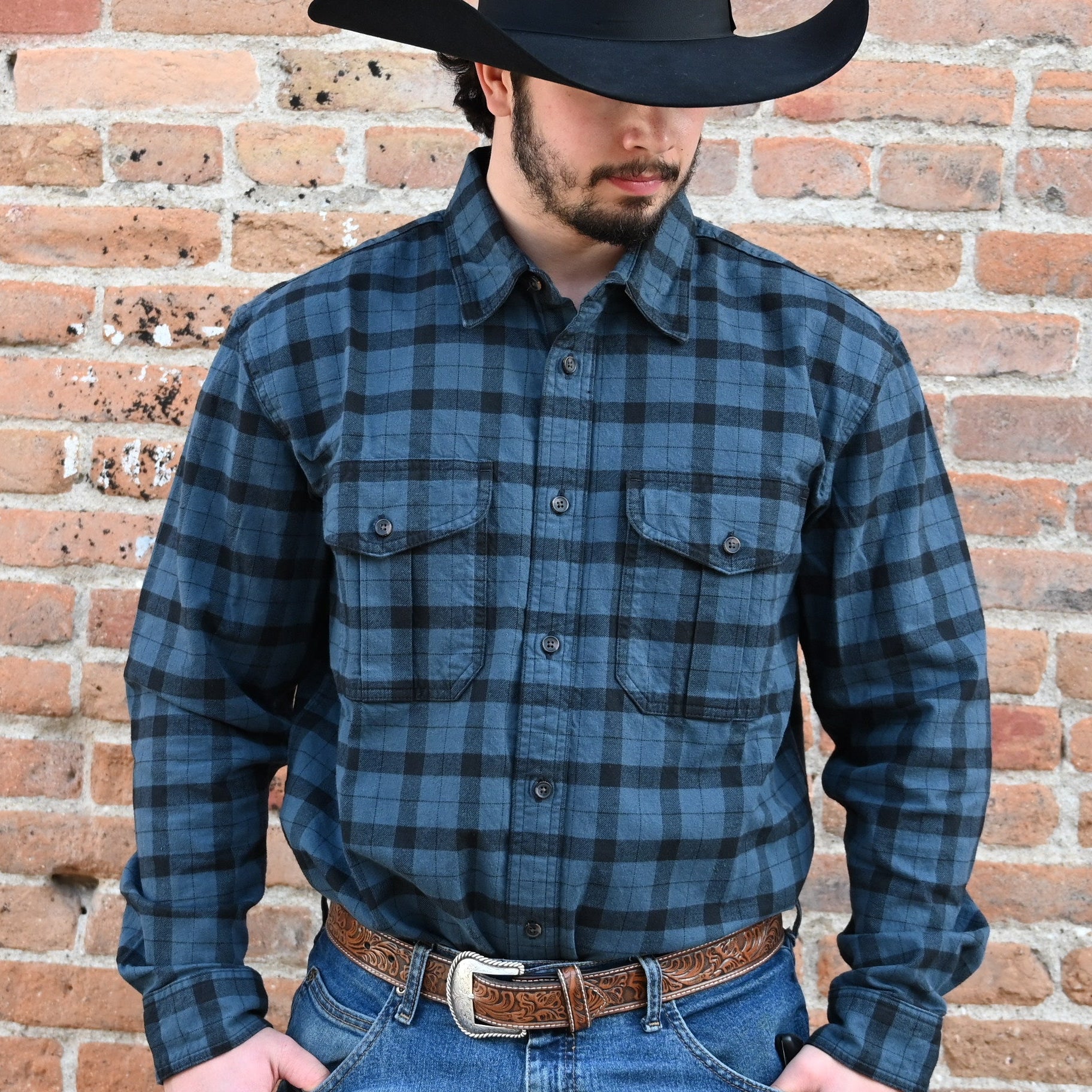 A photo of a man in a plaid shirt and cowboy hat against a brick wall