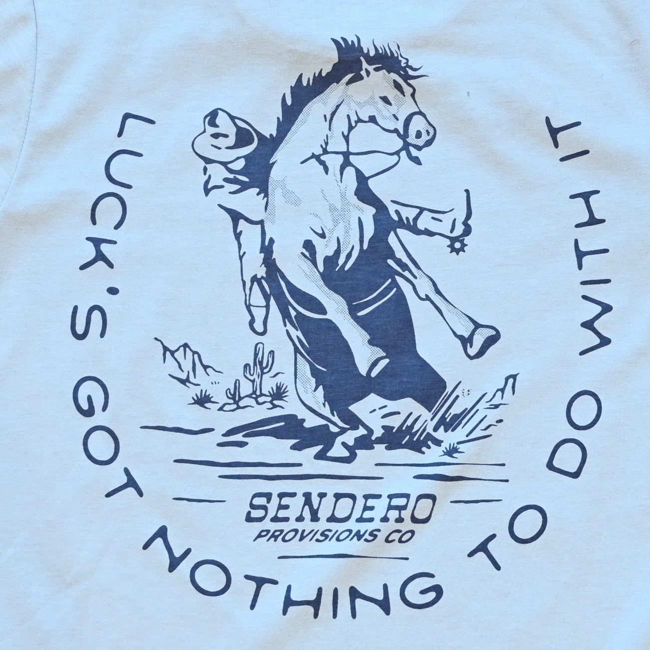 Big Sky Blue No Luck T-Shirt by Sendero with bucking horse graphic and “Luck’s Got Nothin’ to Do With It” text
